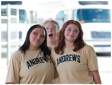 Three adults wearing matching beige shirts with “ANDREWS" printed on them pose together with one person playfully making a surprised expression behind the other two. Description generated by AI