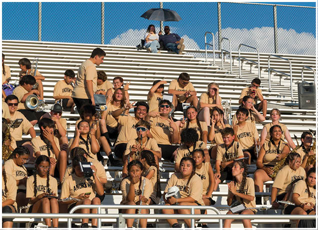 A group of people wearing matching beige shirts are seated on bleachers, some holding musical instruments and others engaging with each other under a clear blue sky. Description generated by AI