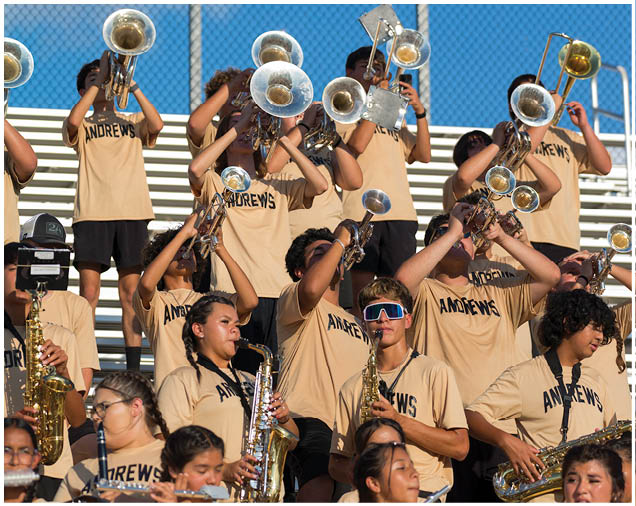 A group of people wearing matching beige shirts with “ANDREWS" printed on them are playing brass and woodwind instruments in a band setting with bleachers and a blue sky in the background. Description generated by AI