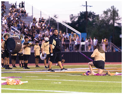 People in casual and uniform clothing are performing on a sports field with musical instruments and flags while a crowd watches from bleachers in the background. Description generated by AI