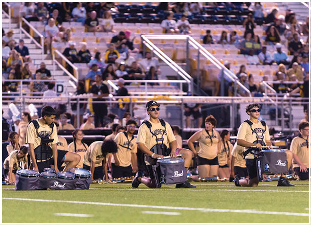Adults wearing matching light-colored shirts and dark shorts are kneeling on a sports field playing drums with a crowd seated in bleachers behind them. Description generated by AI