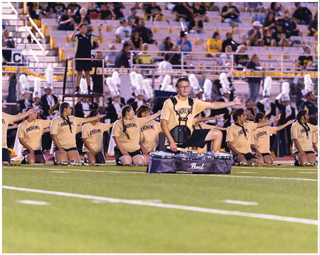 A group of people wearing matching light-colored shirts are kneeling on a field with one person standing behind a drum set in front of them, while an audience watches from the bleachers in the background. Description generated by AI