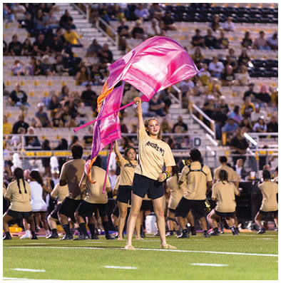 A person wearing a beige Andrews shirt and black shorts is waving a large pink flag on a football field while a marching band performs in the background. Description generated by AI