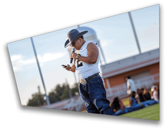 A person wearing a sports jersey and jeans is standing on a field looking at a phone while a group of people sit on the grass in the background near a building. Description generated by AI