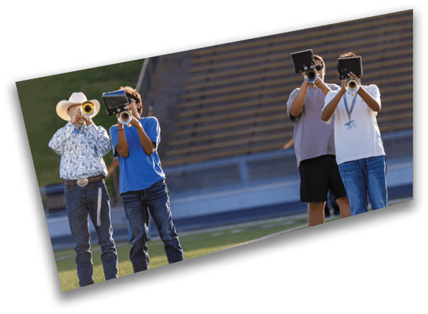 A person in a horse mascot costume is standing on a sports field holding a stick with a horse head on it while a group of people in sports uniforms sit in the background. Description generated by AI