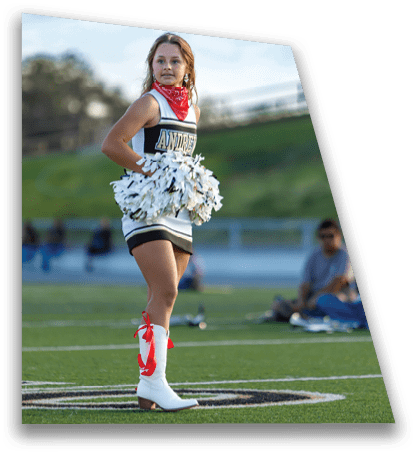 A person wearing a cheerleading uniform with a red bandana and white boots with red ribbons is standing on a sports field holding white pom-poms. Description generated by AI