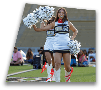 A person in a cheerleading uniform with white boots and red ribbons holds pom-poms on a grassy field with another person kneeling in the background. Description generated by AI
