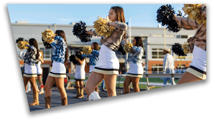 Adults wearing cheerleading outfits and boots are performing with pom-poms on a sports field with a person in a cowboy hat walking in the background. Description generated by AI
