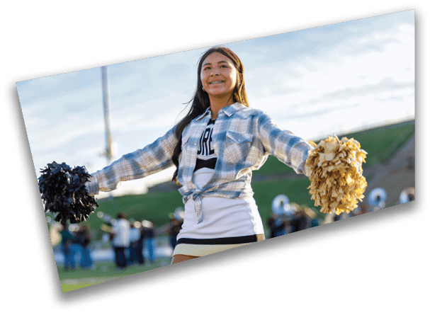 A person wearing a cheerleading outfit with a plaid shirt tied at the waist is holding black and gold pom-poms with arms outstretched on a sports field. Description generated by AI