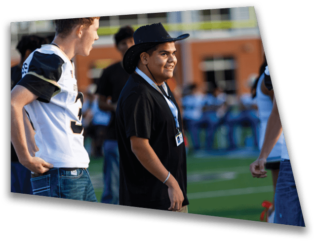A person wearing a black shirt and lanyard is smiling and standing on a sports field near another person in a white sports jersey. Description generated by AI