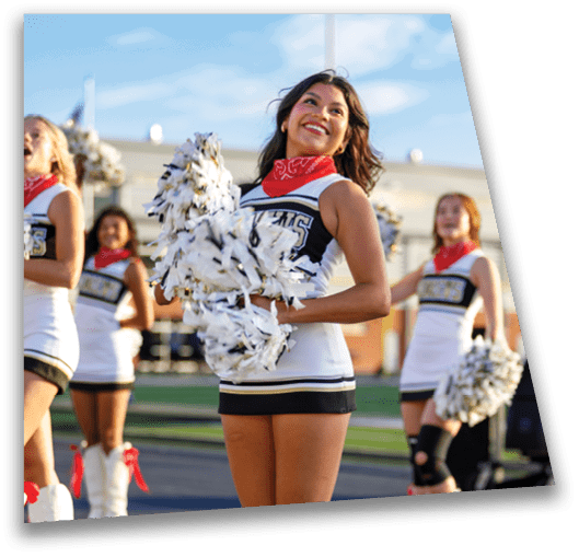 A person wearing a cheerleading outfit and a red bandana around the neck is smiling and holding white pom-poms outdoors. Description generated by AI