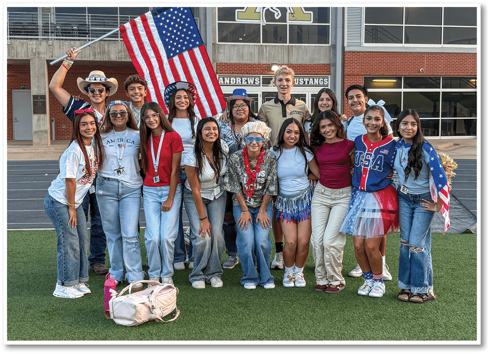 A group of smiling people wearing casual and patriotic clothing pose together outside a building with a large American flag held by one person. Description generated by AI
