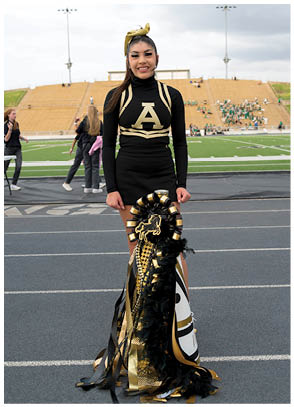 A person wearing a black and gold cheerleading uniform stands on a track in front of a football field holding a decorated black and gold mum. Description generated by AI