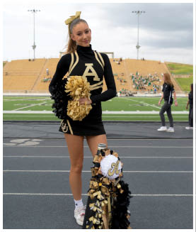 A person wearing a black and gold cheerleading uniform with a large letter A on the front and a matching gold bow in their hair is smiling in front of stadium bleachers under a cloudy sky. Description generated by AI