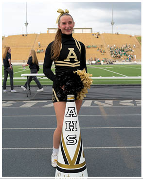 A person in a black and gold cheerleading uniform stands on a track holding black and gold pom-poms behind a white megaphone with the letters AHS in front of a football field and bleachers. Description generated by AI
