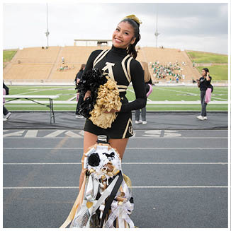 A person in a black and gold cheerleading uniform is smiling and holding black and gold pom-poms on a sports field with a decorated water bottle in front. Description generated by AI