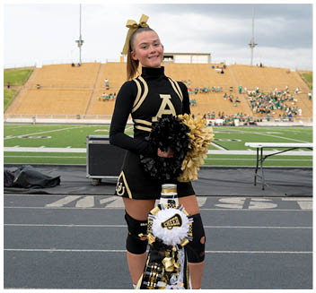A person in a black and gold cheerleading uniform stands on a track field holding black and gold pom-poms with a decorated cheer mum in front and a stadium with spectators in the background. Description generated by AI
