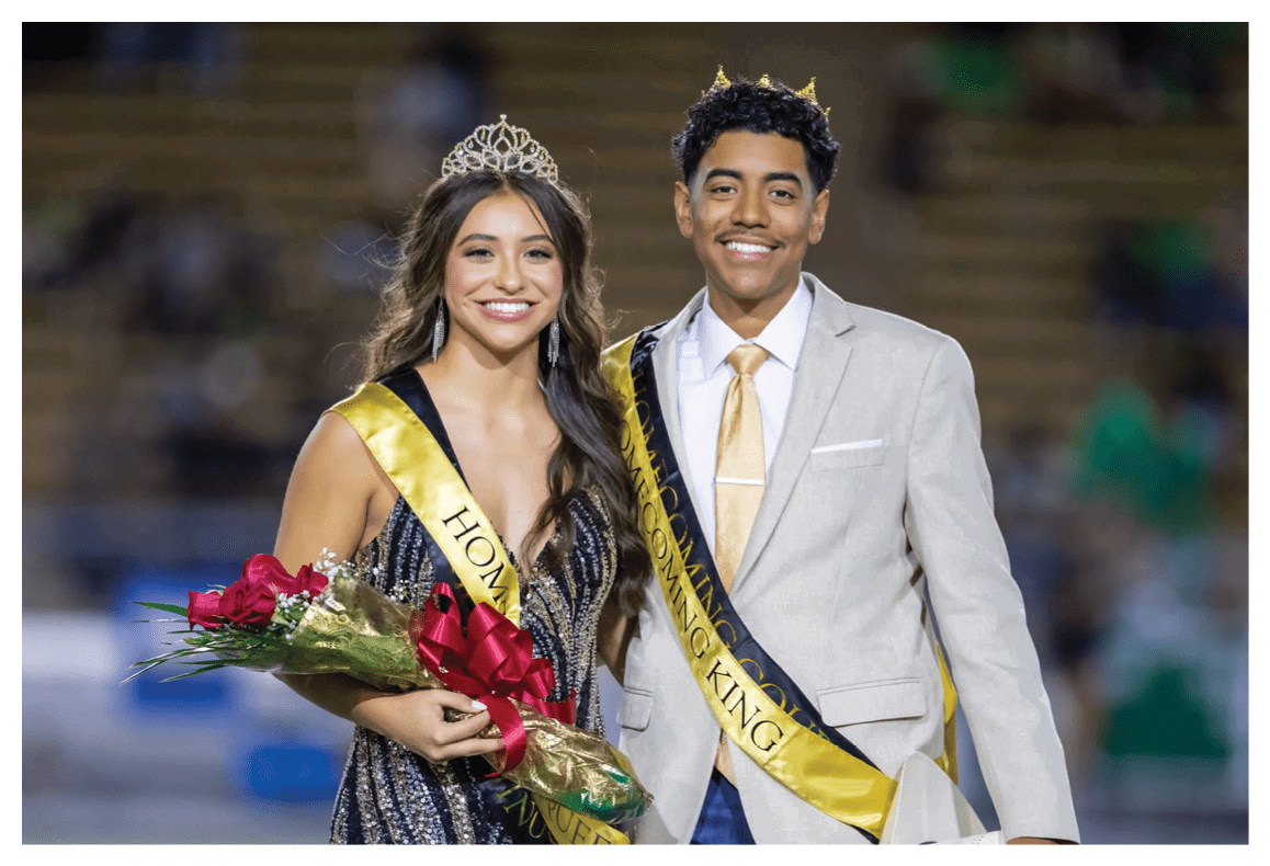 Two people wearing formal attire and sashes labeled Homecoming King and Queen smile while standing together, with one holding a bouquet of flowers. Description generated by AI