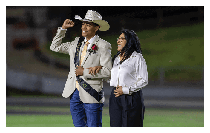 A person wearing a cowboy hat, sash, and boutonniere raises a fist while linking arms with another person dressed in a white blouse and dark pants on a sports field. Description generated by AI