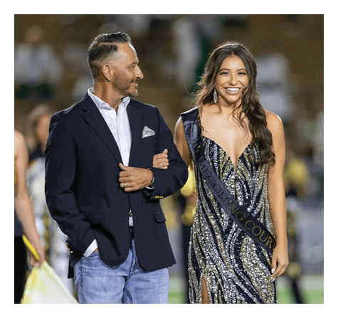A person wearing a sparkly dress and a sash that reads “HOMECOMING COURT" is smiling and walking arm-in-arm with another person dressed in a blazer and jeans at an outdoor event. Description generated by AI