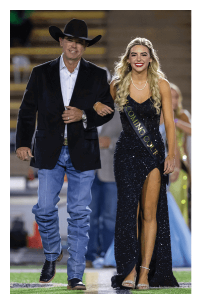 An adult wearing a cowboy hat, blazer, jeans, and boots walks arm-in-arm with another adult in a sparkling black gown with a high slit and a sash that reads “Homecoming Court" on a sports field. Description generated by AI