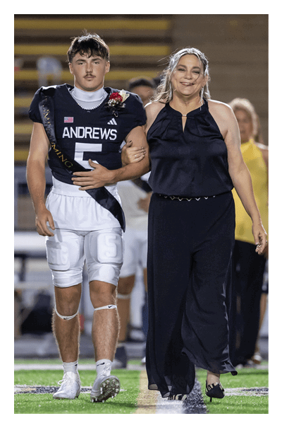 A person in a football uniform with a sash walks arm-in-arm with another person dressed in a black outfit on a sports field with bleachers in the background. Description generated by AI