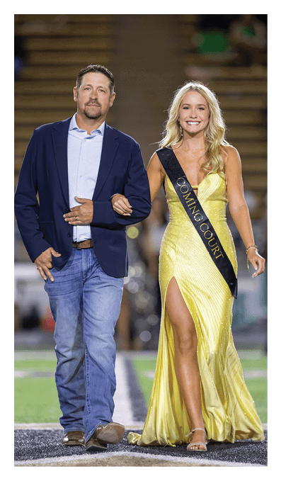 Two adults dressed formally stand side by side with one wearing a dark suit and the other wearing a yellow dress with a sash. Description generated by AI