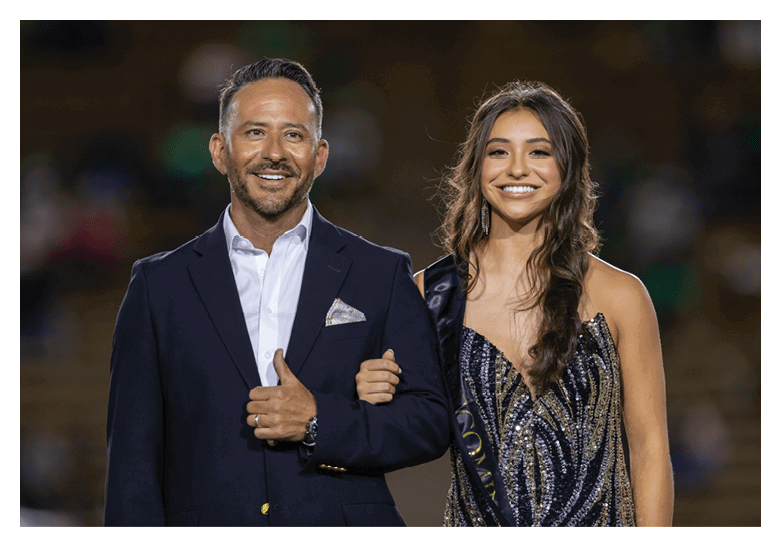 A person in a dark suit with a white shirt and pocket square stands smiling with an arm linked to a person wearing a sparkly dress and a sash, both appearing happy in an outdoor setting. Description generated by AI