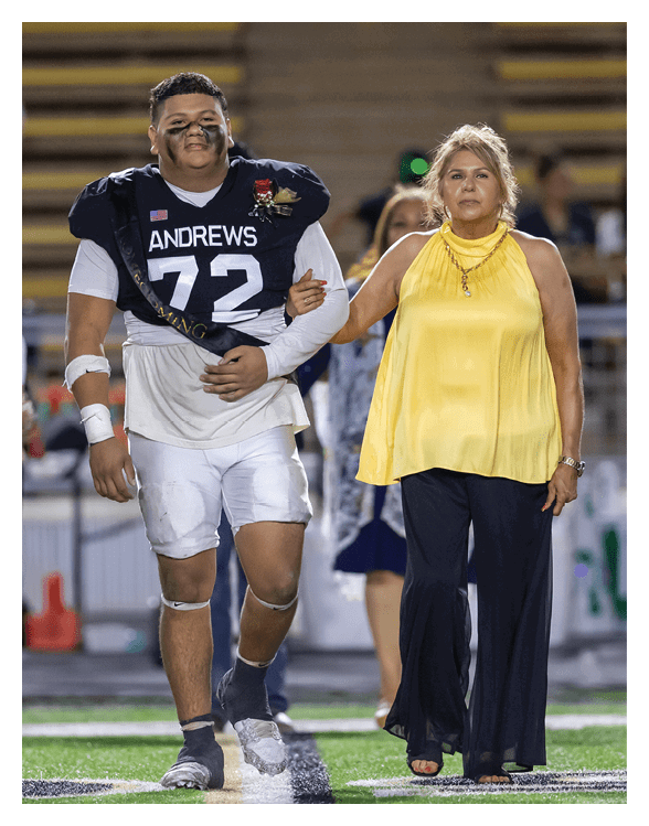 A person wearing a football uniform with the number 72 and a sash walks arm-in-arm with another person dressed in a yellow sleeveless top and black pants on a sports field. Description generated by AI
