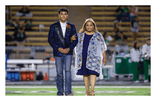 A person wearing a sparkly dark jacket and jeans is walking arm-in-arm with another person in a blue dress and floral shawl on a sports field with bleachers in the background. Description generated by AI