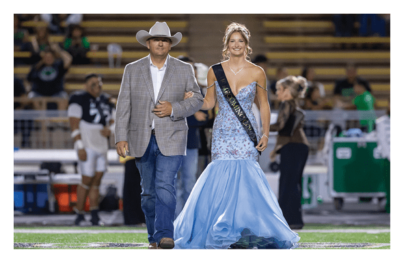 An adult wearing a cowboy hat and blazer walks arm-in-arm with a smiling person in a blue formal gown and sash on a sports field with spectators in the background. Description generated by AI