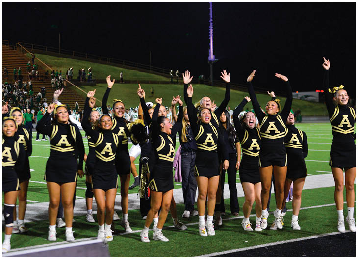 A group of cheerleaders in black and gold uniforms with a large “A" on the front are standing on a football field at night, raising their arms and cheering. Description generated by AI