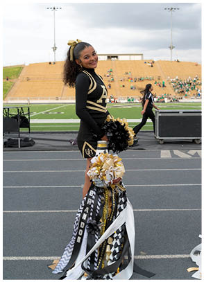 A person in a black and gold cheerleading uniform stands on a track near a football field holding black and gold pom-poms with a large decorated mum in front of them. Description generated by AI