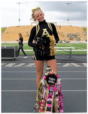 A person in a black and gold cheerleading uniform stands on a track holding black and gold pom-poms with a decorated mum hanging from their waist at a sports stadium. Description generated by AI