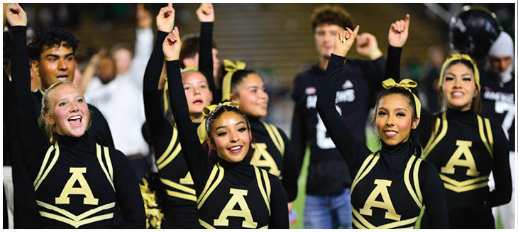 A group of adults in matching black and gold cheerleading uniforms with a large letter A are smiling and raising one arm in a cheer routine. Description generated by AI