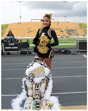 A person in a black and gold cheerleading uniform with a matching bow is standing on a track holding black and gold pom-poms with a decorated mum in front of them and a football field and bleachers in the background. Description generated by AI