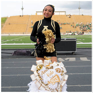 A person in a black and gold cheerleading uniform is smiling and holding black and gold pom-poms while standing behind a large decorated cheer mum on a track near a football field. Description generated by AI