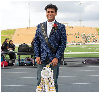 A person wearing a sparkly blue jacket and a sash that says “Homecoming" is standing on a track field holding a decorated homecoming mum with a rose pinned to their jacket. Description generated by AI