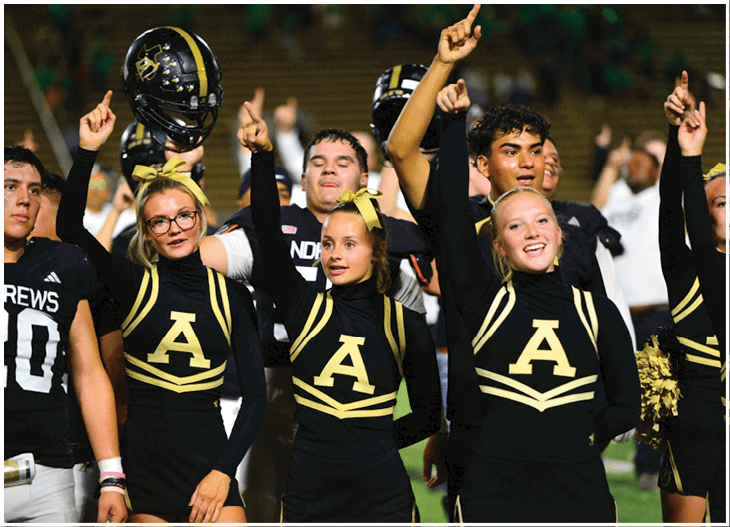 A group of adults in cheerleading uniforms and football gear are raising their index fingers in celebration on a sports field. Description generated by AI