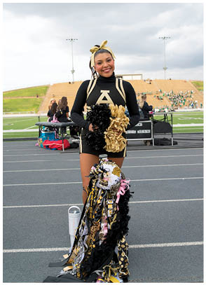 A person wearing a black and gold cheerleading uniform with pom-poms stands on a track in front of a football field holding a large decorated mum. Description generated by AI