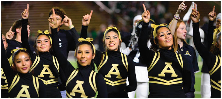 A group of adults in black and gold cheerleading uniforms with large letter A on the front are raising one finger in the air, smiling, and standing closely together at an outdoor event. Description generated by AI