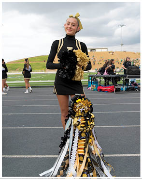 A person in a black and gold cheerleading uniform stands on a track holding black and gold pom-poms with a large decorative mum adorned with ribbons and trinkets in front of them at a sports field. Description generated by AI