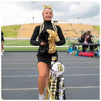 A person in a black and gold cheerleading uniform stands on a track holding black and gold pom-poms with a decorated homecoming mum attached to their leg in a stadium setting. Description generated by AI