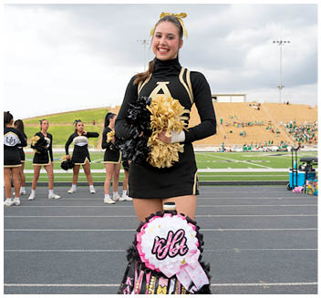 A person in a black and gold cheerleading uniform stands on a track holding black and gold pom-poms with a decorated mum hanging in front, while other cheerleaders stand in the background near a football field. Description generated by AI