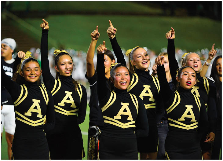 A group of adults in matching black and gold cheerleading uniforms with large letter A on the front are smiling and raising one arm with a finger pointed upward on a sports field. Description generated by AI