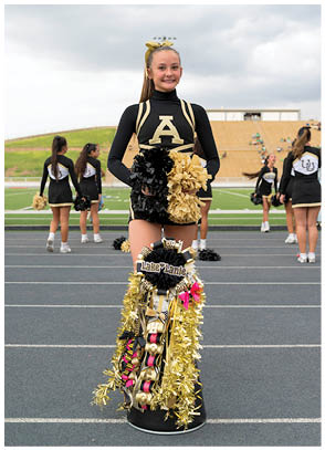 A person in a black and gold cheerleading uniform stands on a track holding black and gold pom-poms with a decorated black and gold mum hanging in front, while other cheerleaders stand in the background on a football field. Description generated by AI