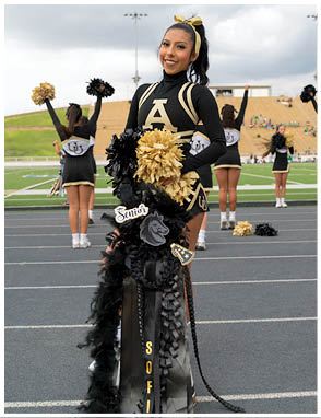 A person in a black and gold cheerleading uniform stands on a track holding pom-poms and decorated items with the word “Senior" while other cheerleaders perform in the background on a sports field. Description generated by AI