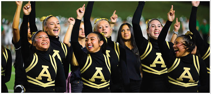 A group of adults in matching black and gold cheerleading uniforms with the letter A are smiling and raising their arms enthusiastically. Description generated by AI
