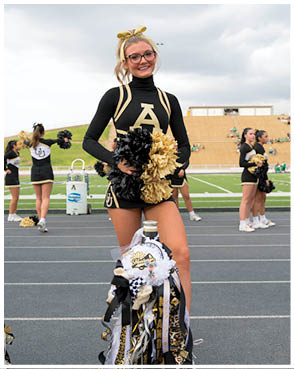A person wearing a black and gold cheerleading uniform with a large letter A on the front is standing on a track holding black and gold pom-poms with a decorated mum hanging in front. Description generated by AI
