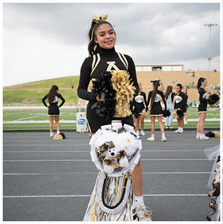 A person in a black and gold cheerleading uniform stands on a track holding black and gold pom-poms with a decorated white and gold mum hanging from their waist while other cheerleaders stand in the background near a football field. Description generated by AI